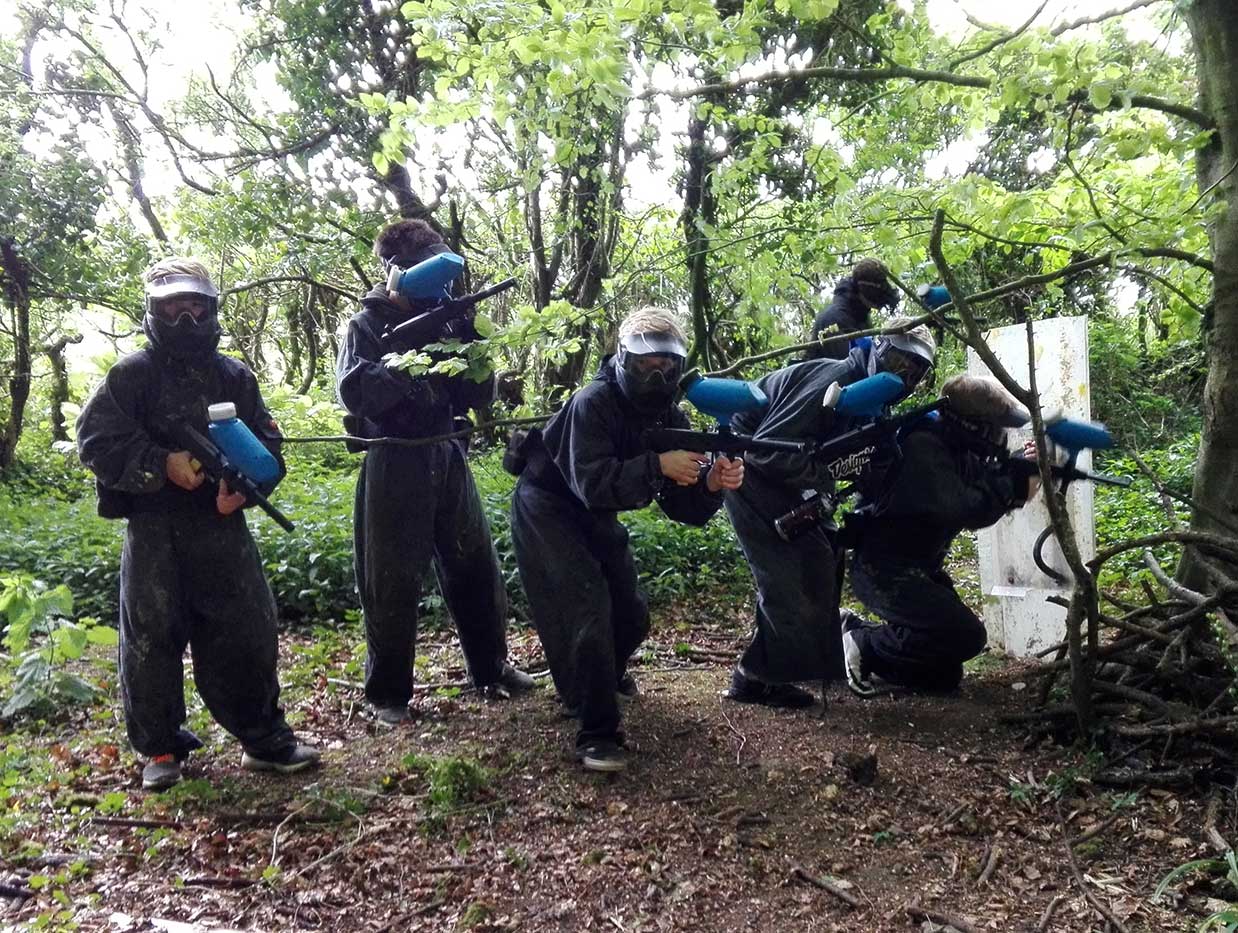 A squad of kids armed with paintball guns and a riot shield attacks up a hill.
