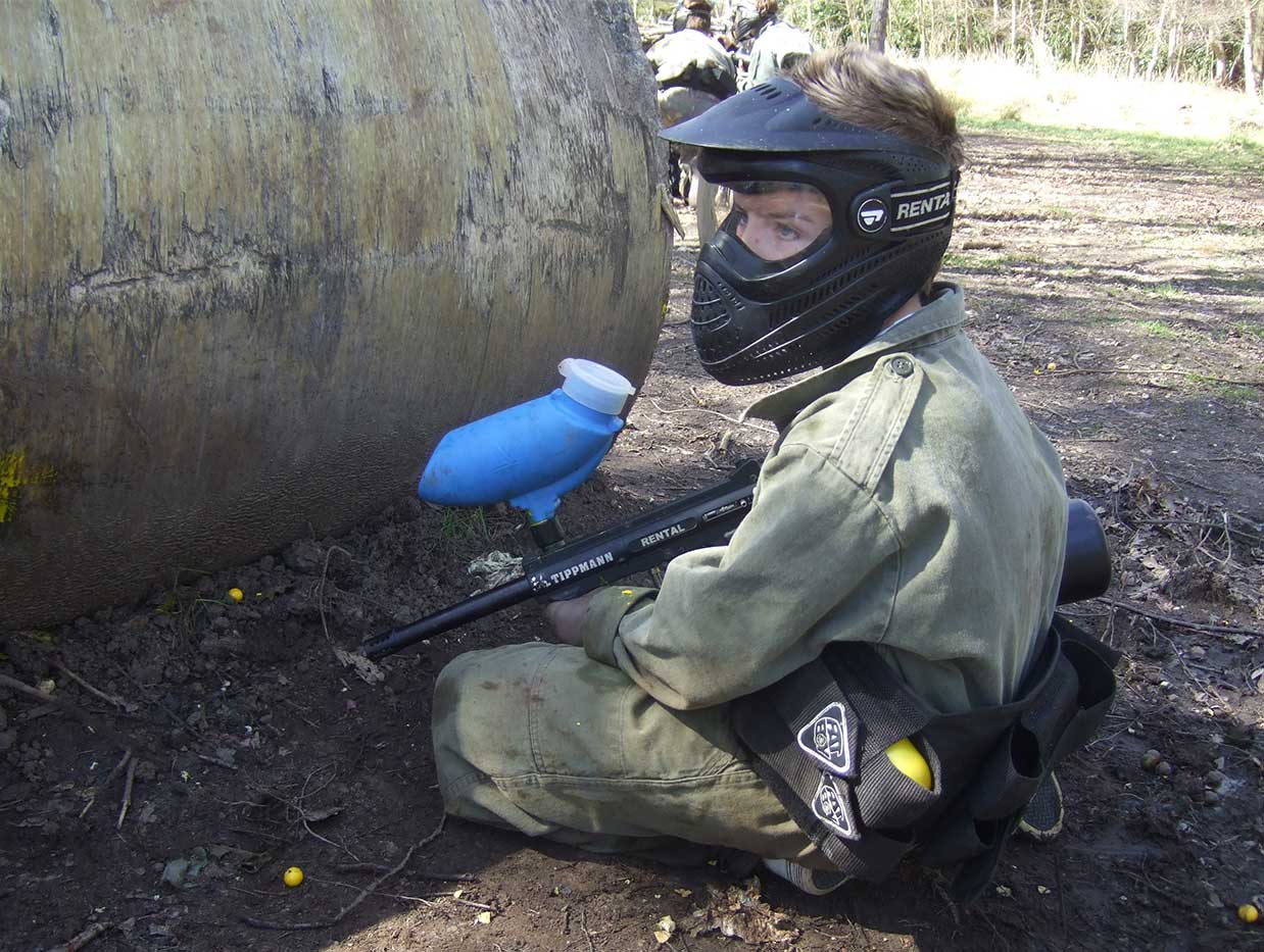 Kid resting behind a large pipe with his gun at the ready during a paintball game.